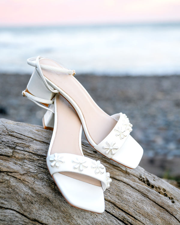White high-heeled shoes with floral details on a wooden log by a body of water.