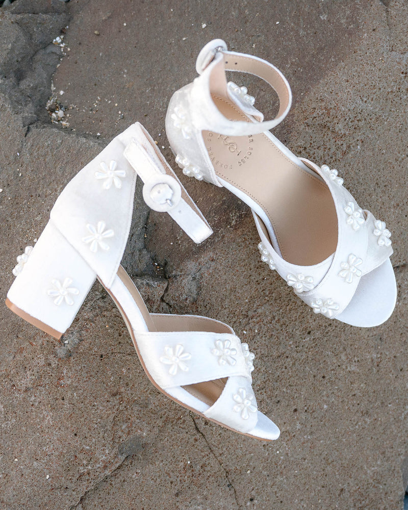 White high-heeled sandals with floral details on a sandy surface