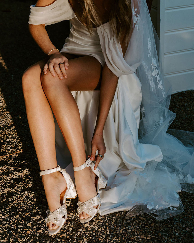 Person wearing a white dress and bridal high heels sitting on a gravel surface.