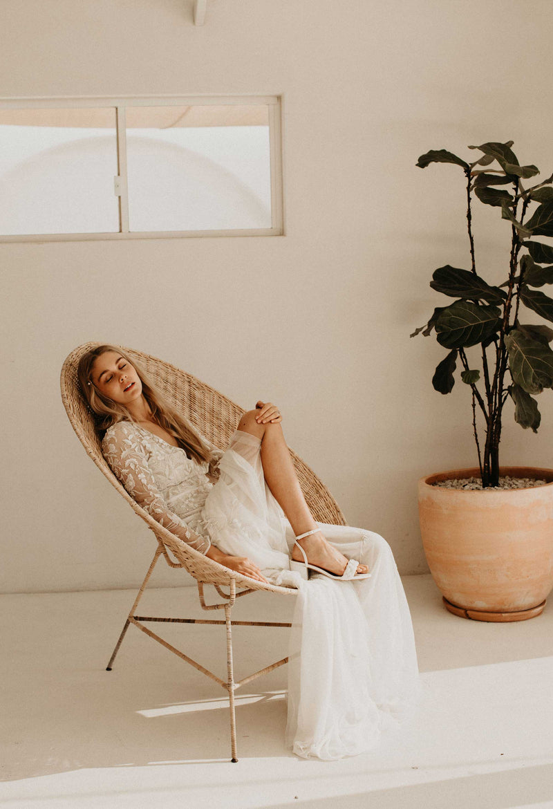 Woman in a white dress sitting in a wicker chair in a minimalistic room with a plant.