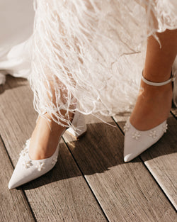 White wedding shoes with floral details worn by a person in a white dress on a wooden floor.