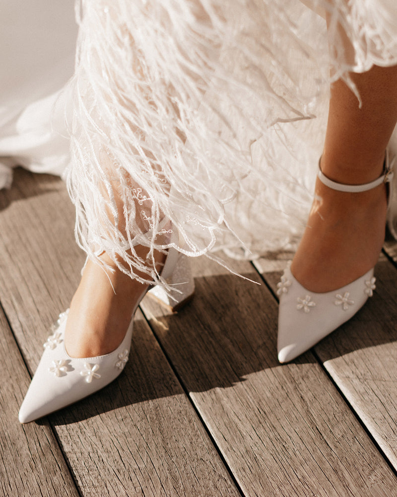 White wedding shoes with floral details worn by a person in a white dress on a wooden floor.