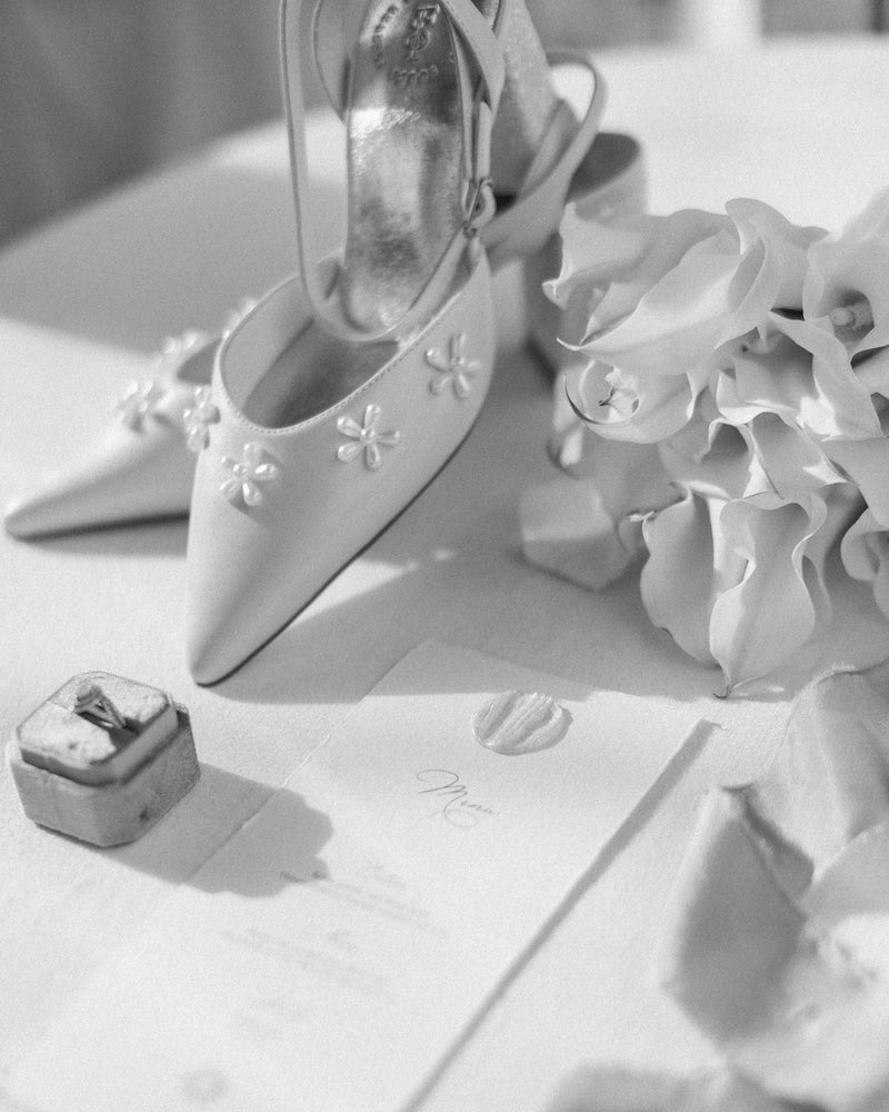 Black and white image of wedding shoes with a ring box and bouquet on a table.