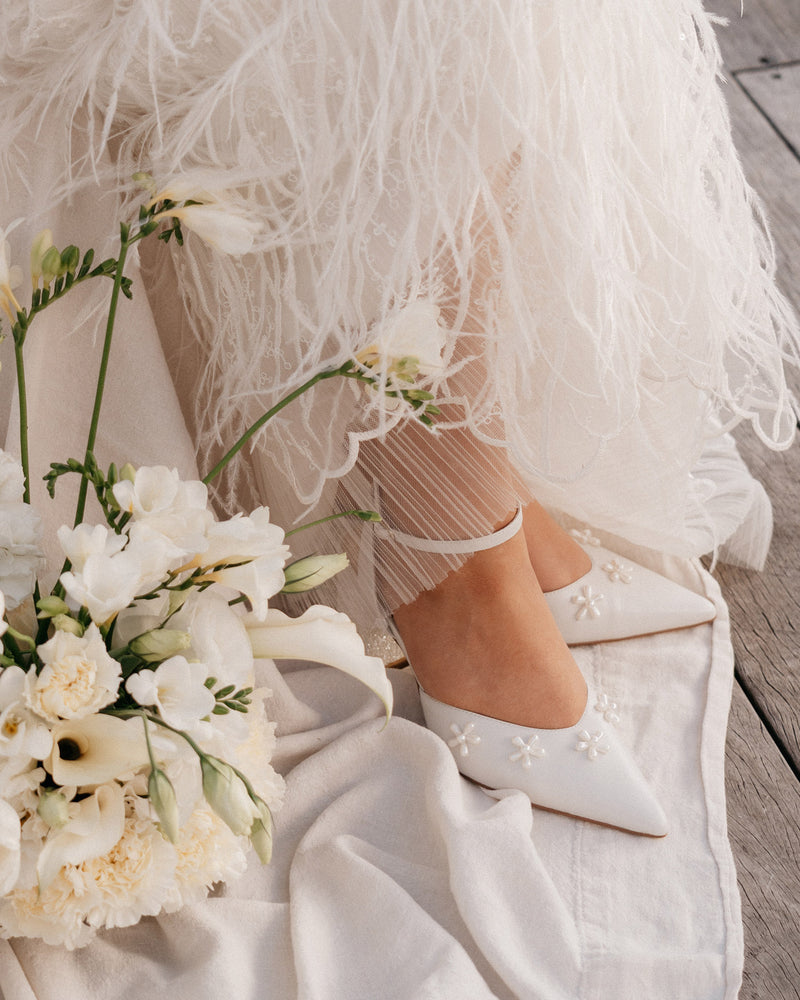 White wedding shoes with floral details worn by a bride, sitting on a wooden floor with a bouquet of white flowers.