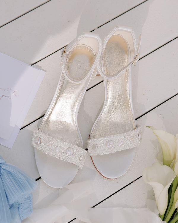 White wedding shoes with bead details on a wooden floor with flowers and a dress.