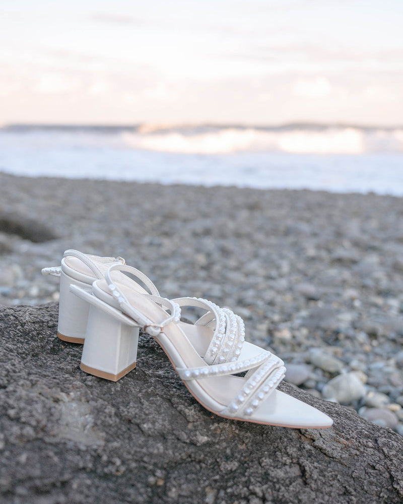 White high-heeled sandals with pearl details on a pebbly surface near water.