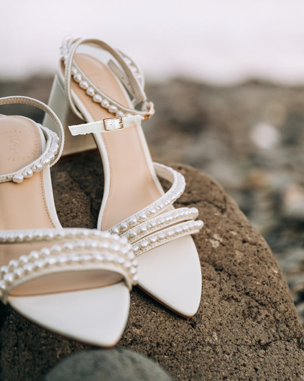 Beige high-heeled sandals with pearl embellishments on a rocky surface.